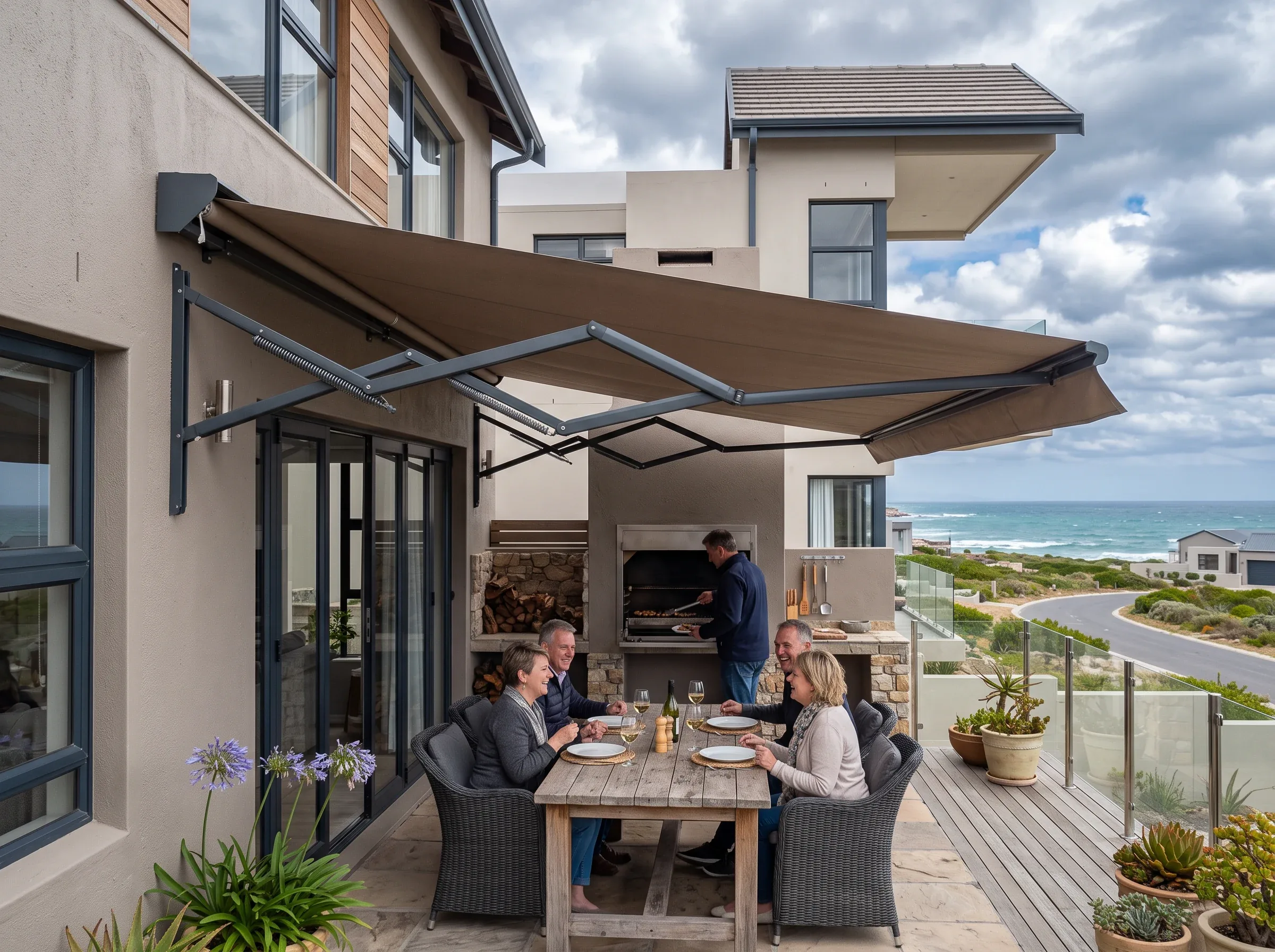 Folding-arm awning extended from the wall above a built-in braai, four people at a weathered timber table under it, glass balustrade and a coastal street with breaking surf beyond, heavy cloud overhead